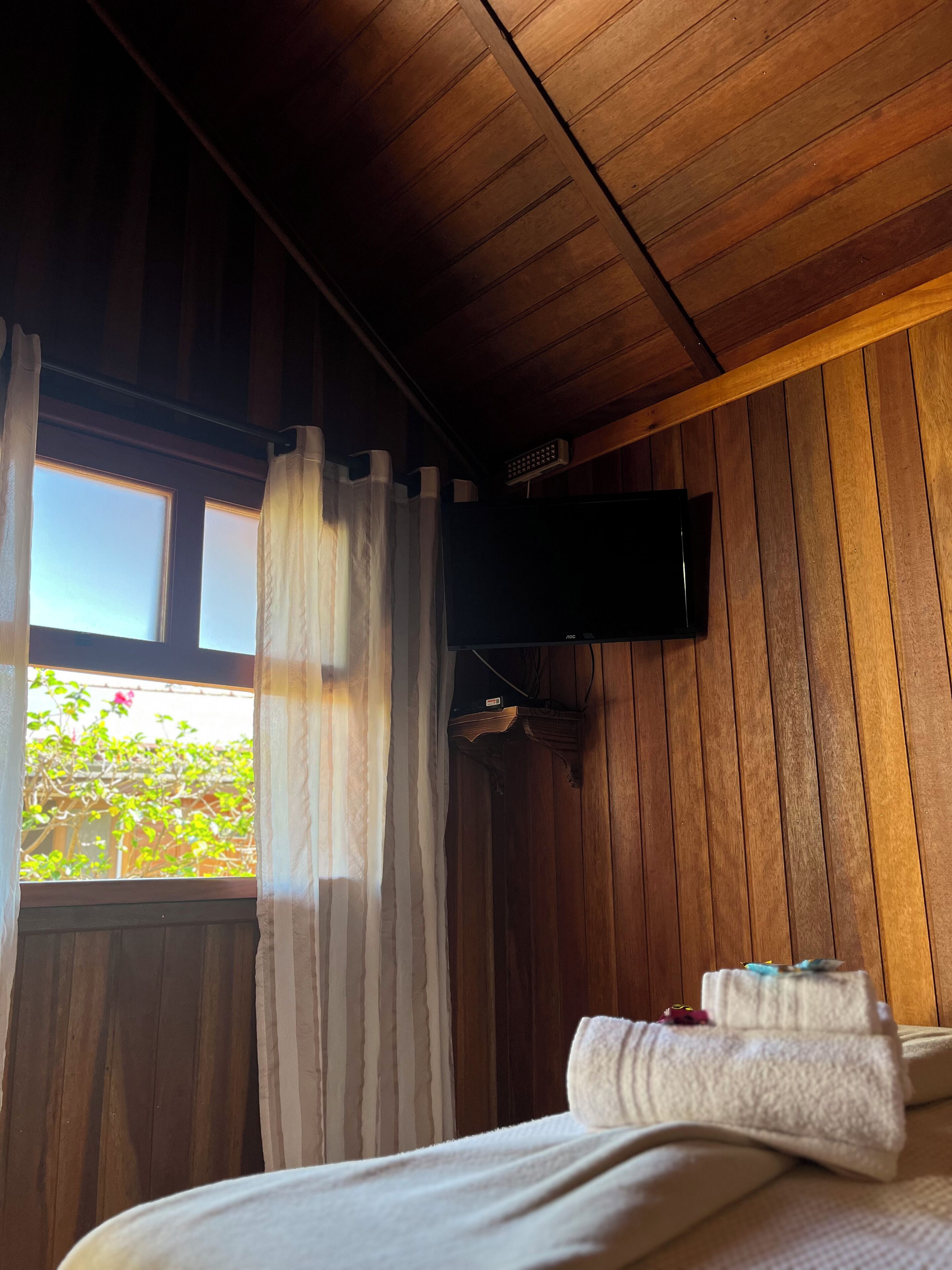 Una habitación con paneles de madera, una cama, un televisor montado en la pared y una ventana con cortinas.