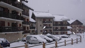 Parcheggio innevato con diverse auto e edifici sullo sfondo.