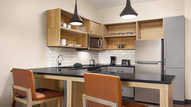 A modern kitchen with wooden cabinets, a black countertop, two orange chairs, and stainless steel appliances. - Candlewood Suites Panama City Tyndall AFB by IHG (Panama City)