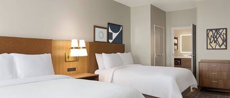Ein Hotelzimmer mit zwei Betten, einem Holzkopfstück, einem Nachttisch mit Lampe, einem Schminktisch und einer Tür zu einem Badezimmer.