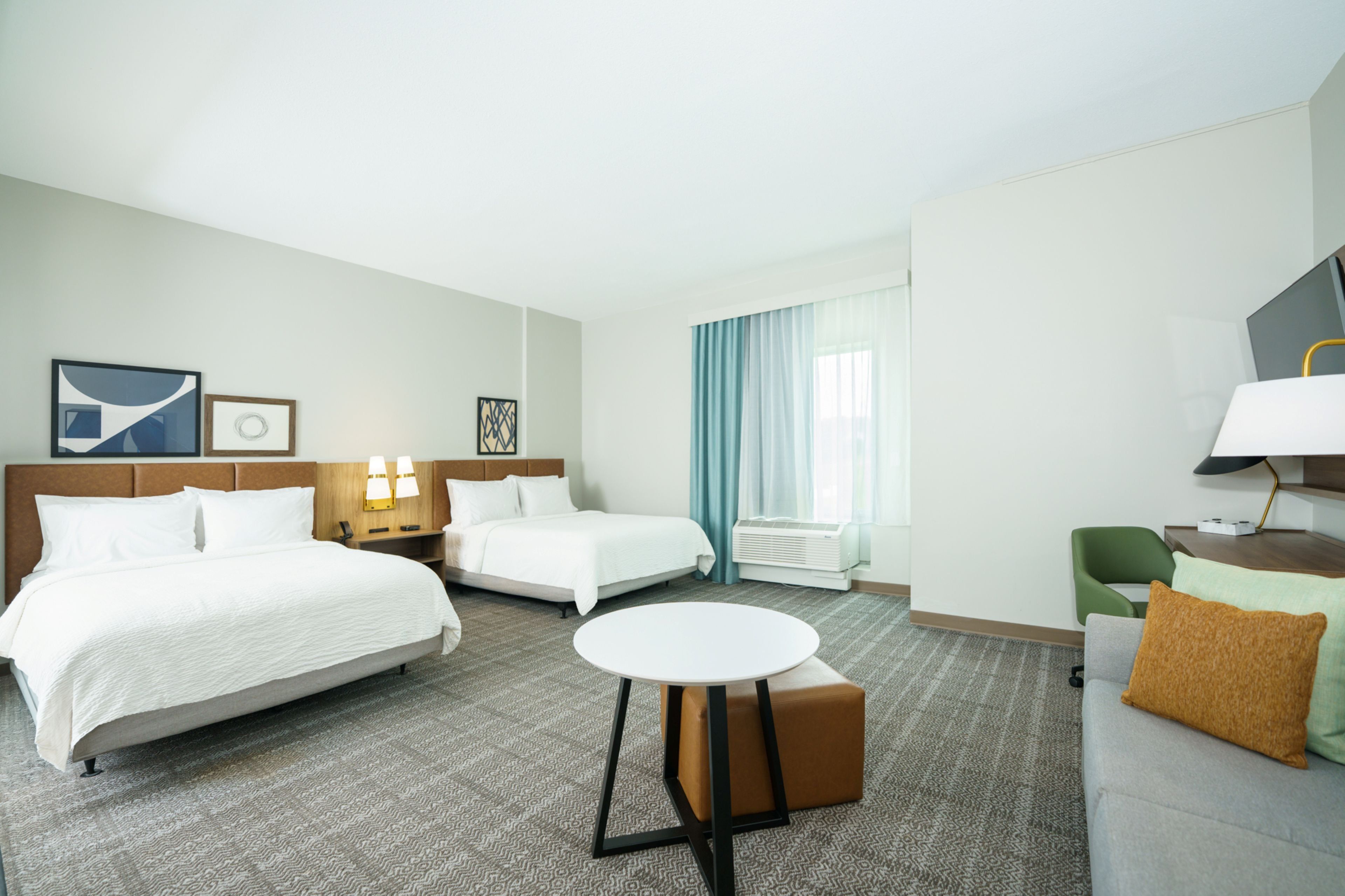 Une chambre d’hôtel avec deux lits, une table ronde, une chaise verte, un téléviseur et une fenêtre avec des rideaux.