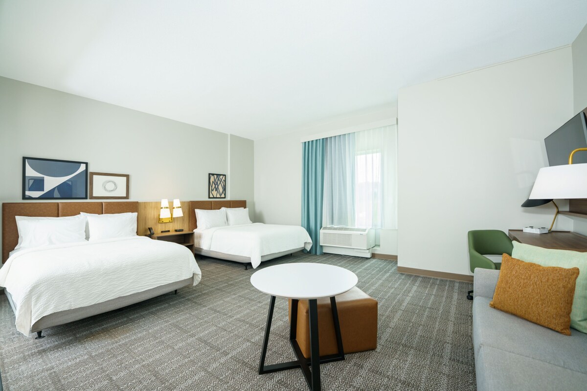 A hotel room with two beds, a round table, a green chair, a television, and a window with curtains.