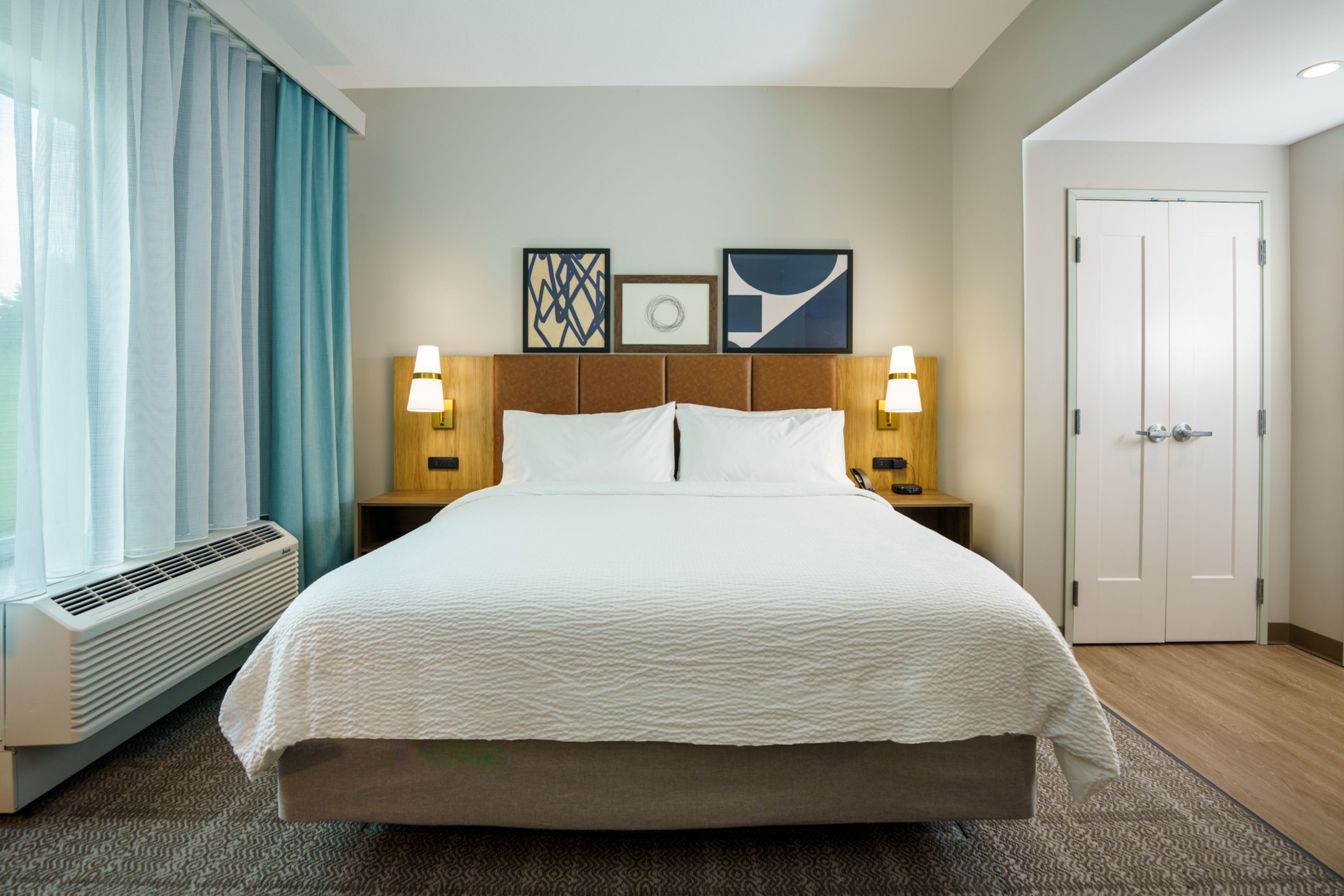 Une chambre d’hôtel avec un grand lit, deux tables de chevet avec des lampes, une fenêtre avec des rideaux et une porte.
