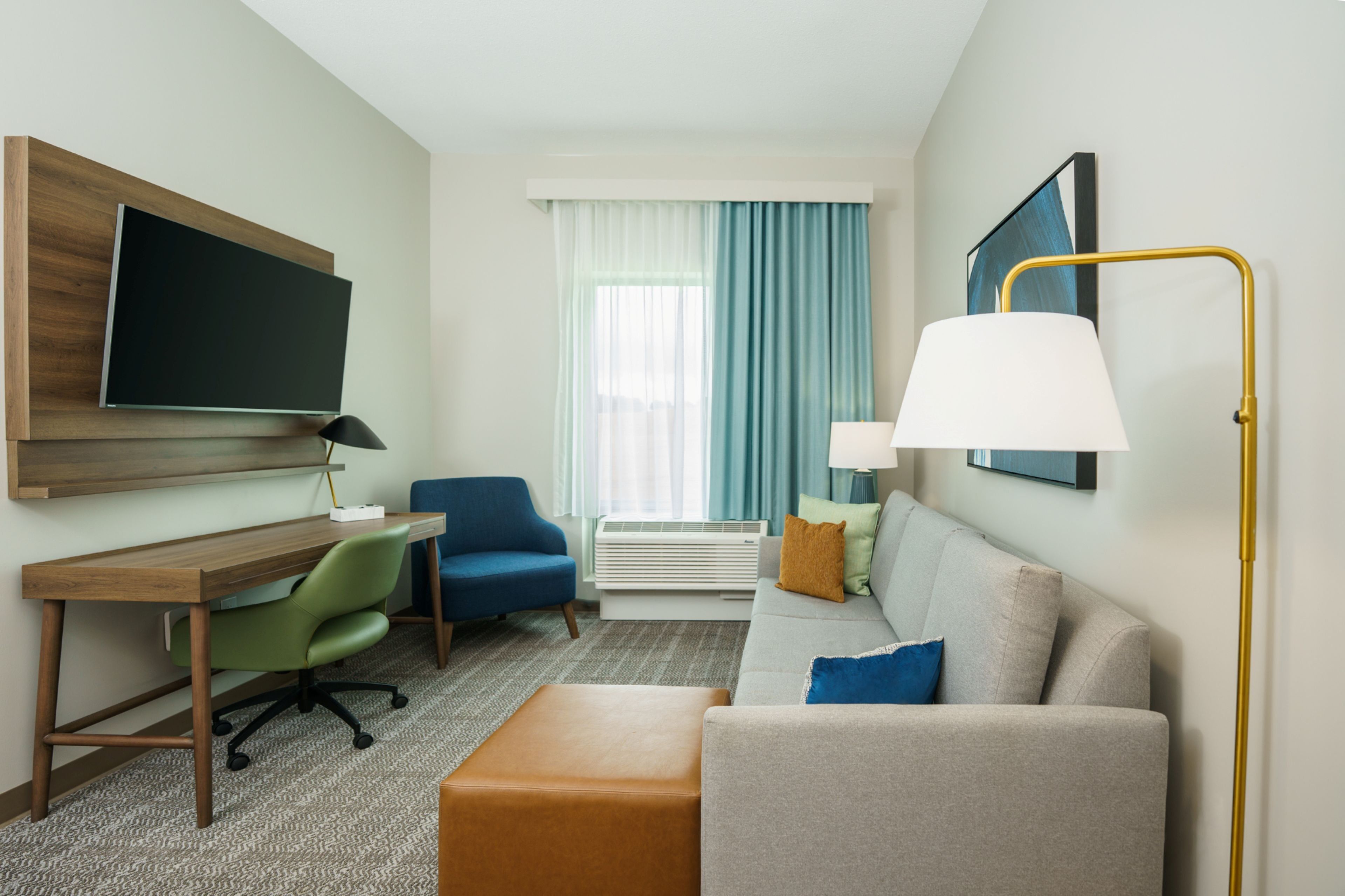 Habitación de hotel con escritorio, una silla azul, sofá con cojines, lámpara de pie y televisor de pantalla plana montado en la pared.