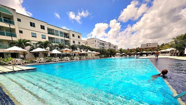 A swimming pool with lounge chairs and umbrellas, surrounded by a building with multiple floors and balconies. - Palm Village (Ipojuca)