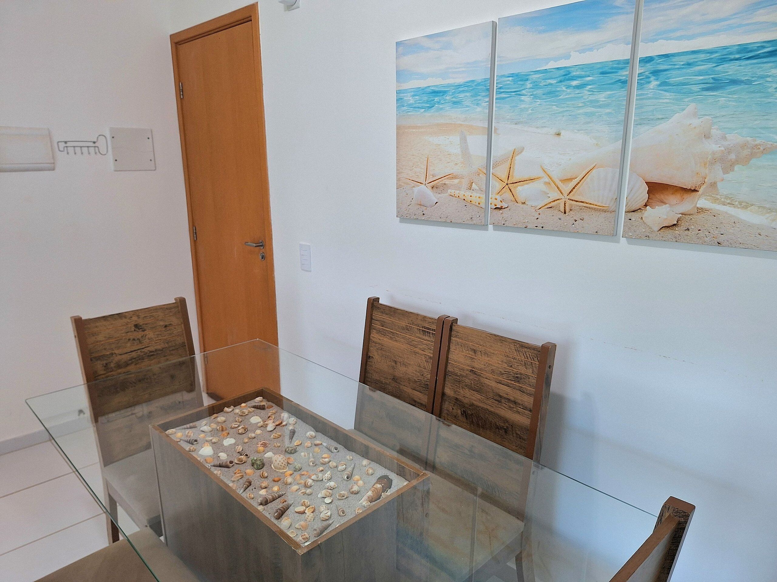 Una habitación con una mesa de comedor de vidrio rodeada de sillas de madera, una puerta de madera y un cuadro colgado en la pared que representa una escena de playa.