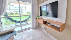A modern living room with a large TV, a hammock, and a view of a green outdoor area. - Palm Village (Ipojuca)