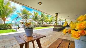 An outdoor seating area with wooden tables and benches, surrounded by palm trees and a potted plant with yellow flowers. - Palm Village (Ipojuca)