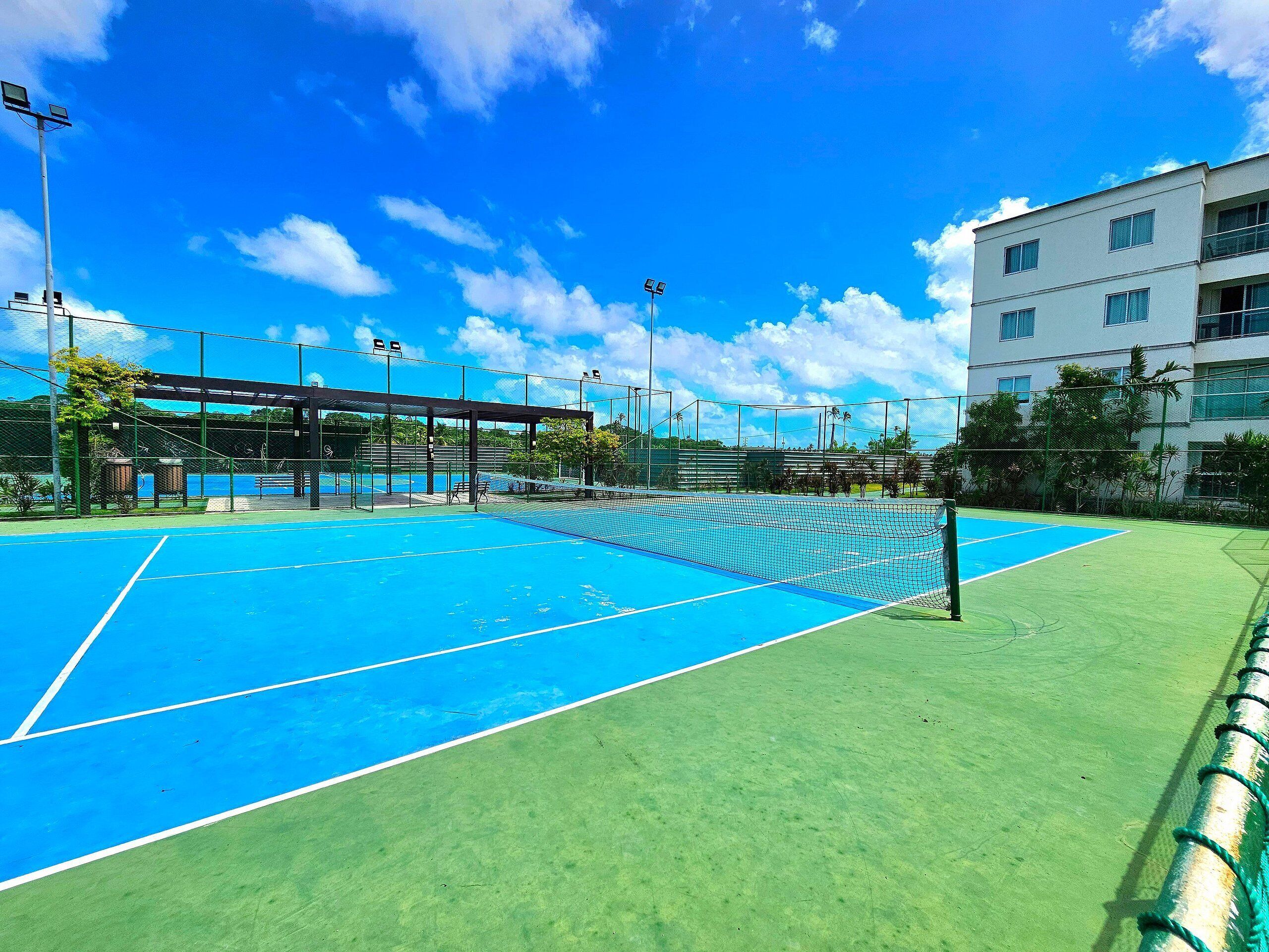 An outdoor tennis court with a net, surrounded by a fence and a covered seating area.