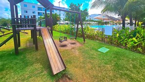 A playground with a slide and swings, surrounded by grass and palm trees, with a pool and umbrellas in the background. - Palm Village (Ipojuca)