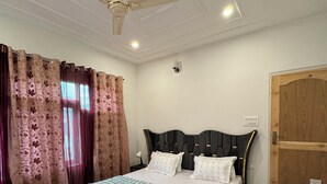 A bedroom with a bed, curtains, a ceiling fan, a wooden door, and a nightstand. - Shesh Bagh Boutique Homestay (Srinagar)