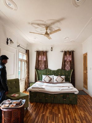 A bedroom with a green upholstered bed, a ceiling fan, patterned curtains, and a wooden floor. - Shesh Bagh Boutique Homestay (Srinagar)