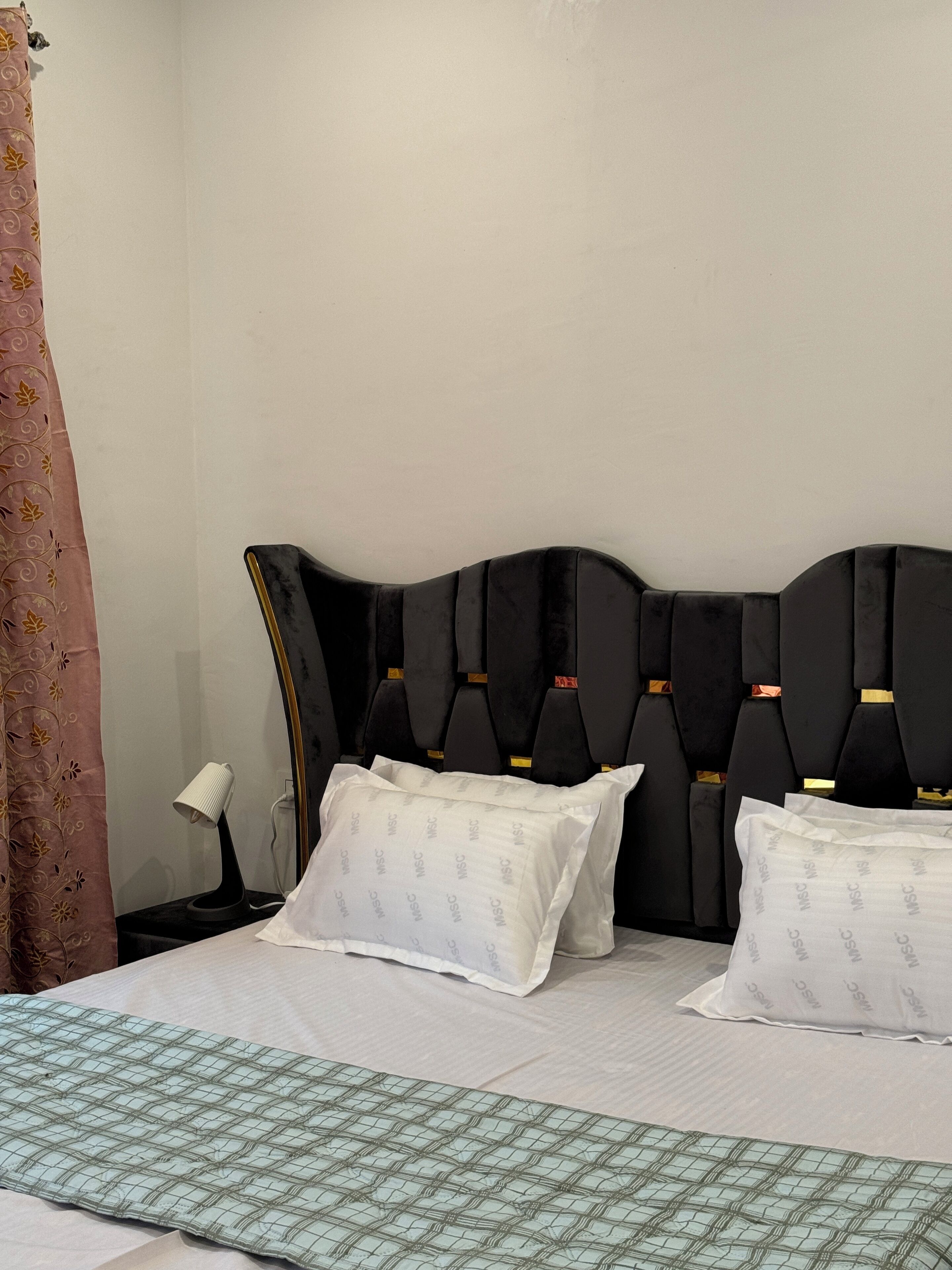 A bed with a black headboard, two white pillows, a green patterned bedspread, and a nightstand with a lamp.