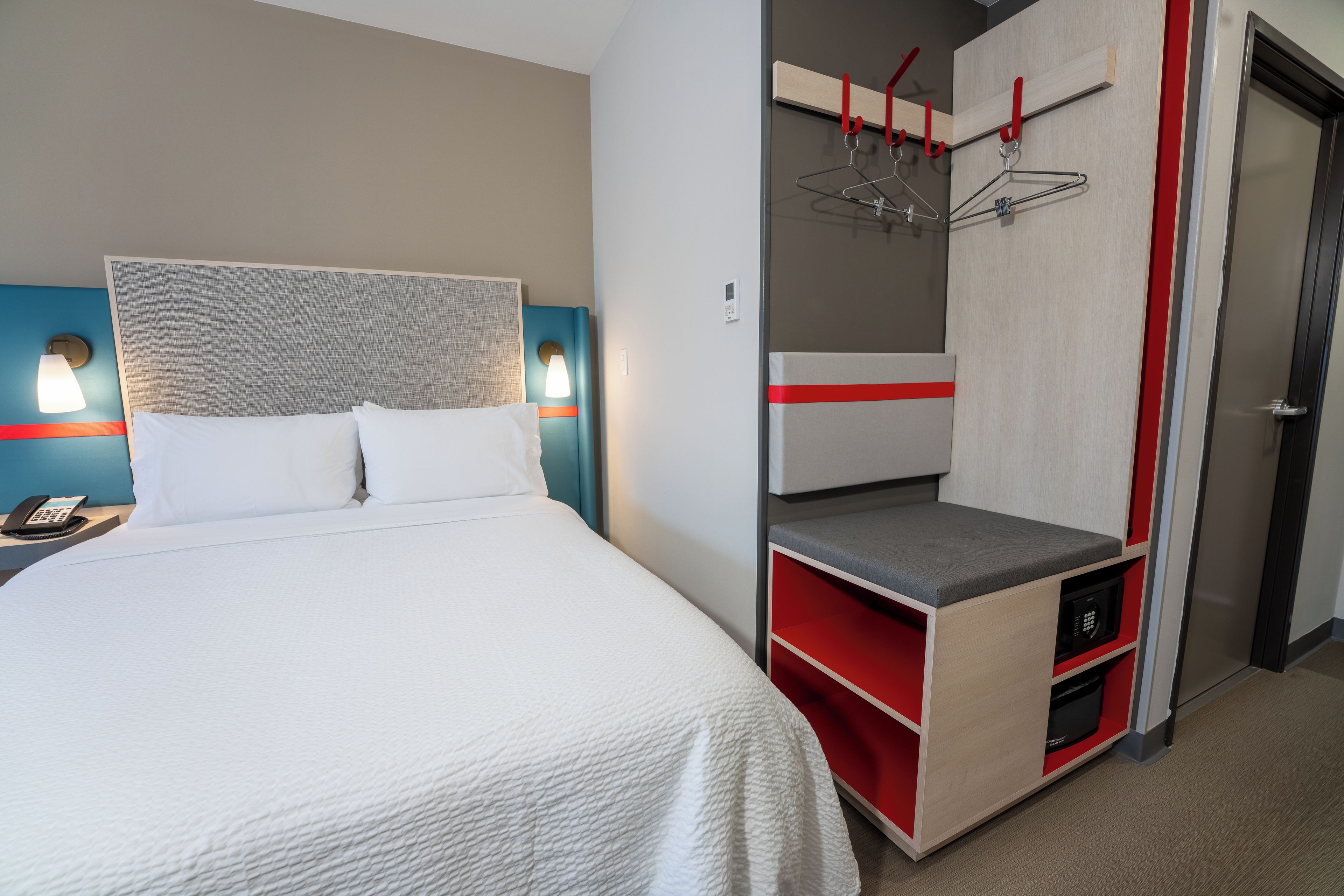 Una habitación de hotel con cama, escritorio con teléfono y armario con detalle rojo.