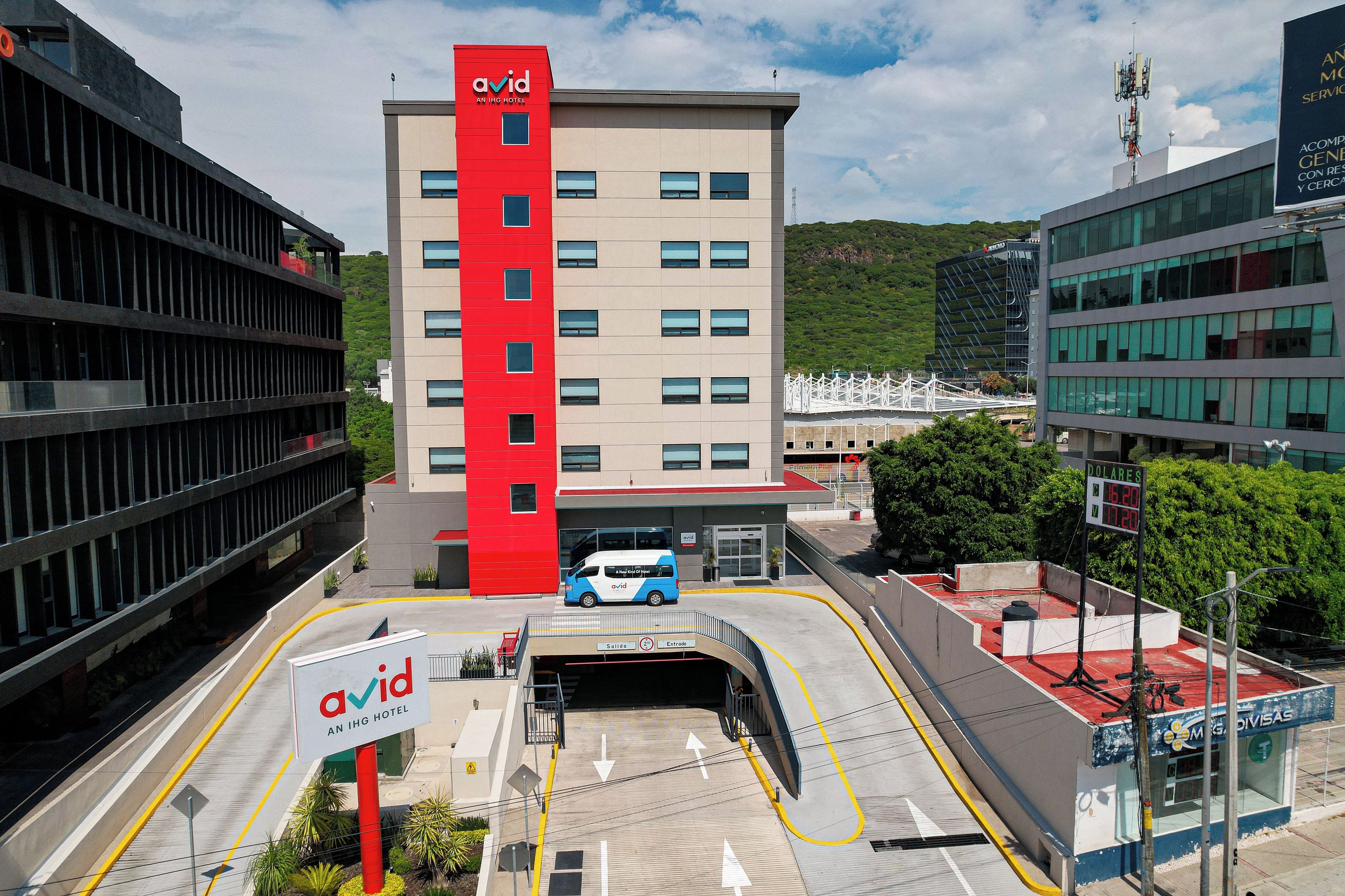 A modern hotel with a red and beige facade, a parking area, and a view of a green hill in the background.