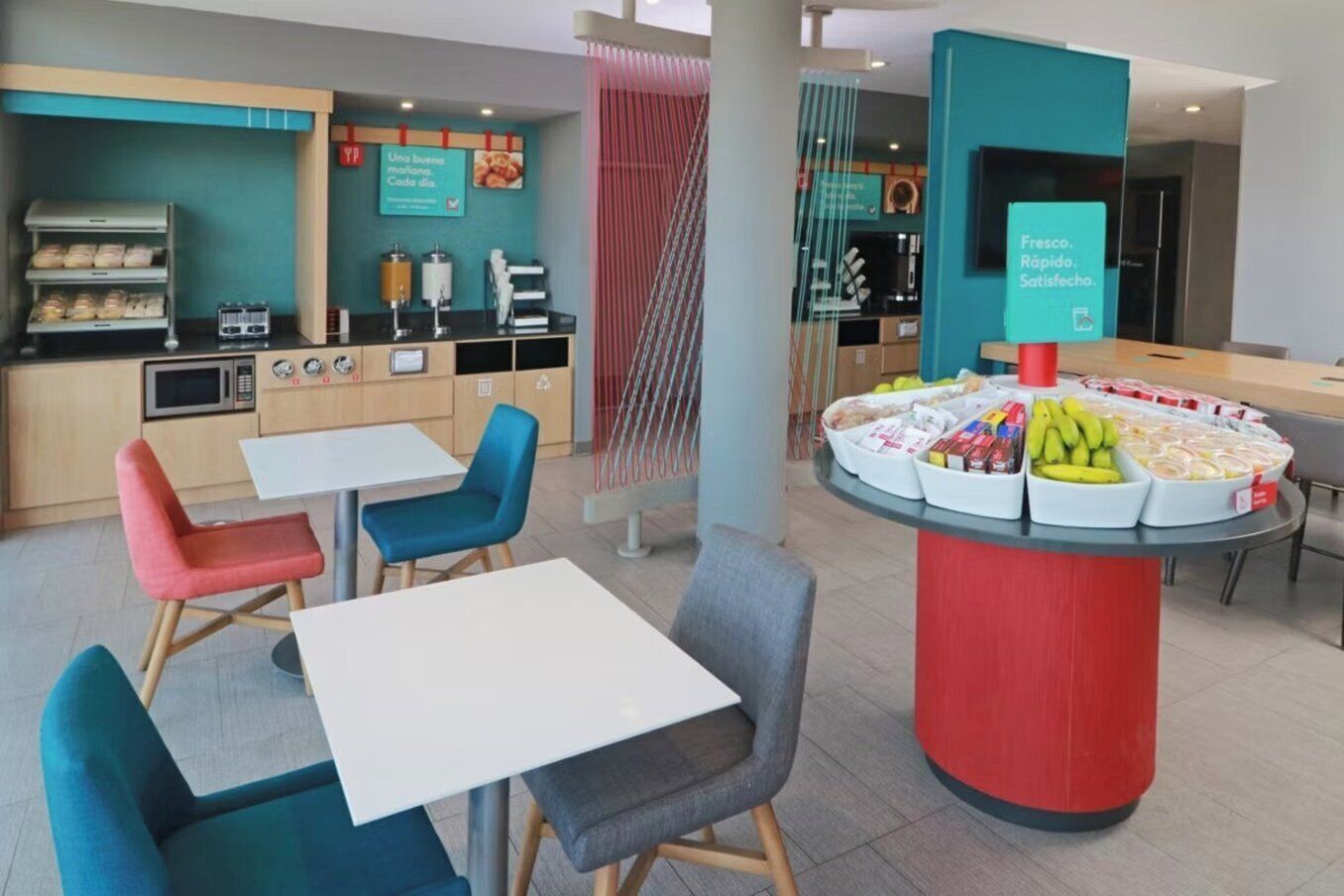 Un café moderno con una mesa redonda central que exhibe frutas frescas y aperitivos, rodeado de sillas coloridas y una zona de cocina al fondo.