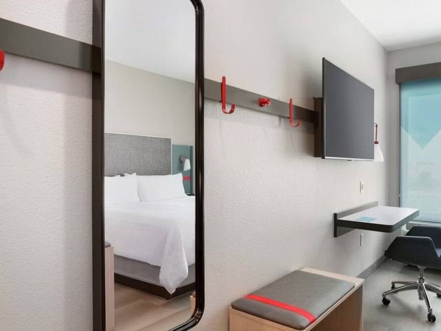 Una habitación de hotel moderna con cama, escritorio, silla, televisor y espejo.