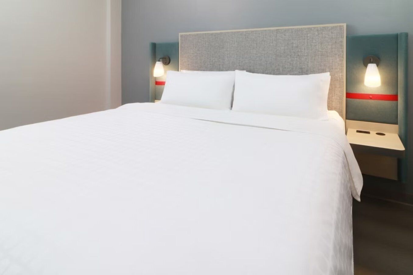 A neatly made bed with white linens, two bedside tables with red accents, and wall-mounted lamps.
