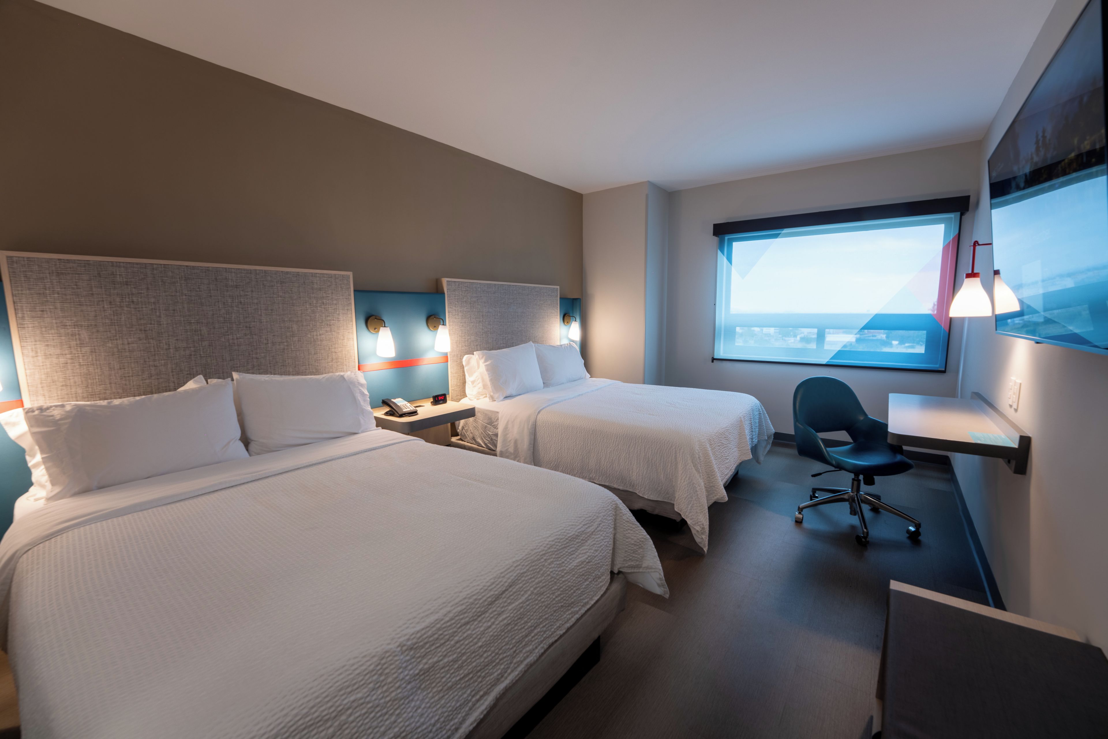 A hotel room with two beds, a desk, a chair, and a large window offering a city view.