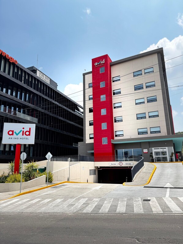Edificio de hotel moderno con fachada roja y gris, un letrero que indica que es un hotel IHG y una entrada despejada con una línea amarilla de seguridad.