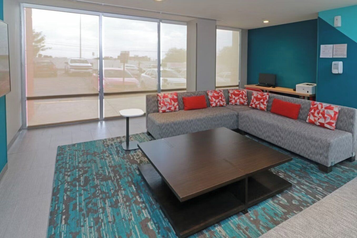 A modern living room with a grey sectional sofa, red patterned cushions, a dark wooden coffee table, and a teal patterned rug.
