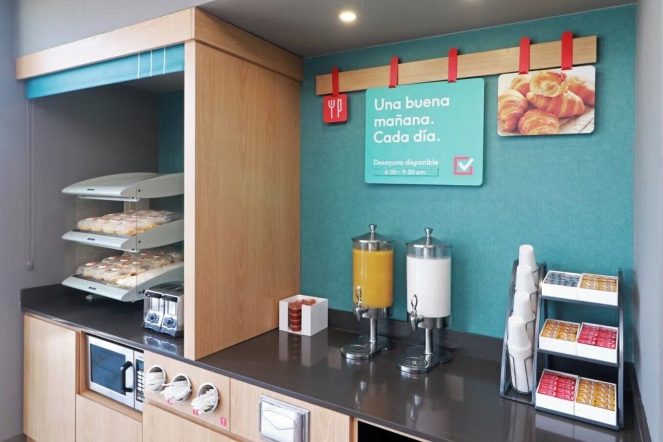 A breakfast area with a self-service station, including a microwave, juice dispensers, and a variety of packaged foods.