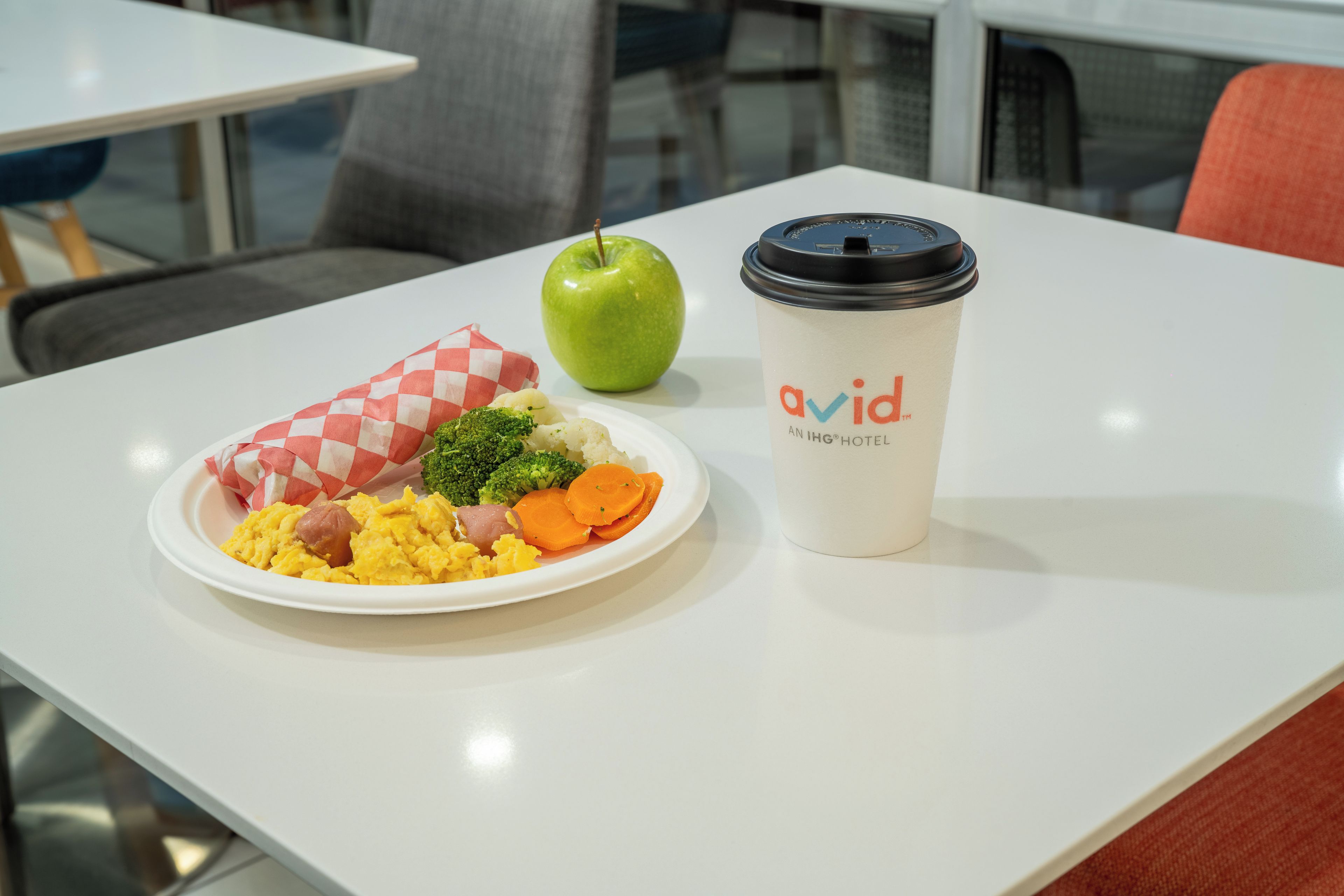Una mesa blanca con un desayuno que incluye huevos revueltos, salchicha, brócoli y zanahorias, además de una taza de café con tapa.