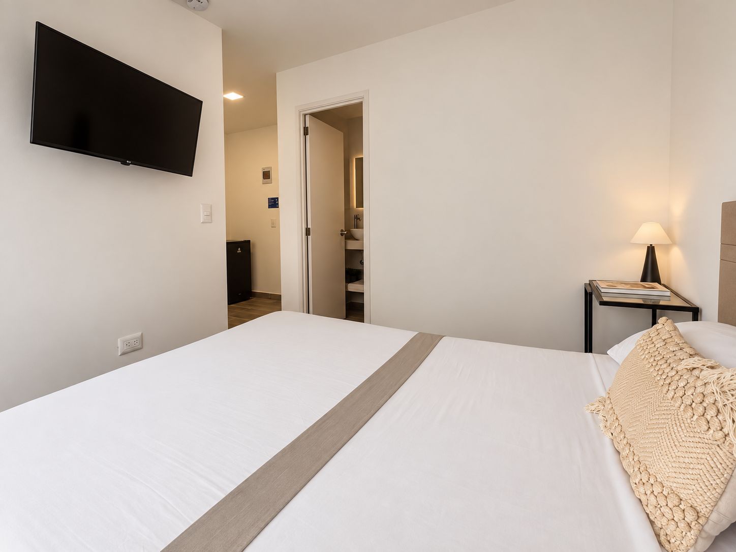 Habitación de hotel con cama, televisión montada en la pared, mesita de noche con lámpara y puerta que da a un baño.
