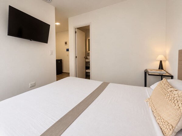 A hotel room with a bed, a television mounted on the wall, a bedside table with a lamp, and a door leading to a bathroom. - Apartasuites ISABEL (Cali)