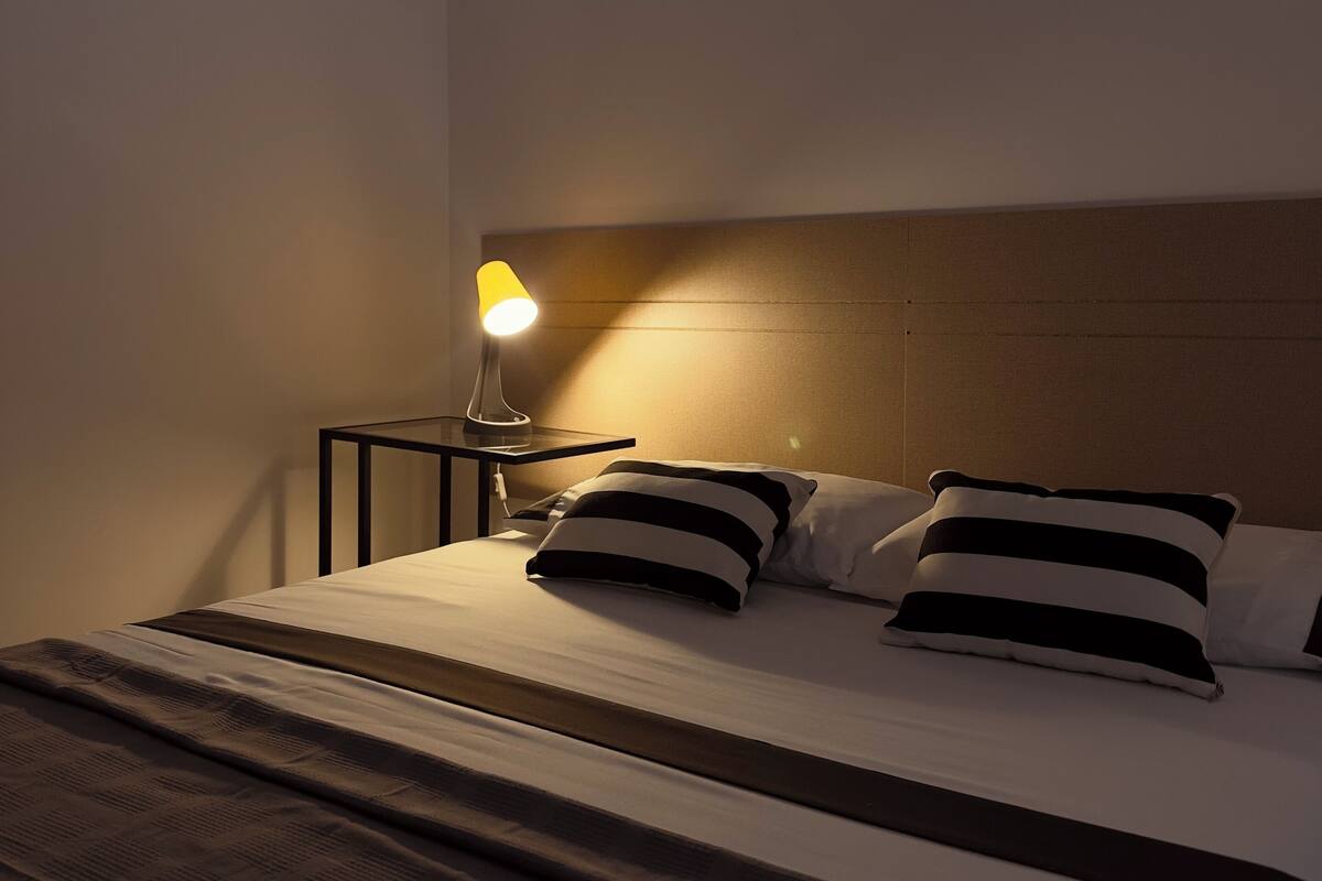 A neatly made bed with striped pillows, a bedside table with a lamp, and a wooden headboard.