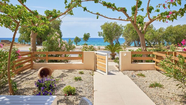 A pathway leading to a beach with a wooden fence and a gate, surrounded by greenery and flowers. - Votsalo Ierapetra (Ierapetra)