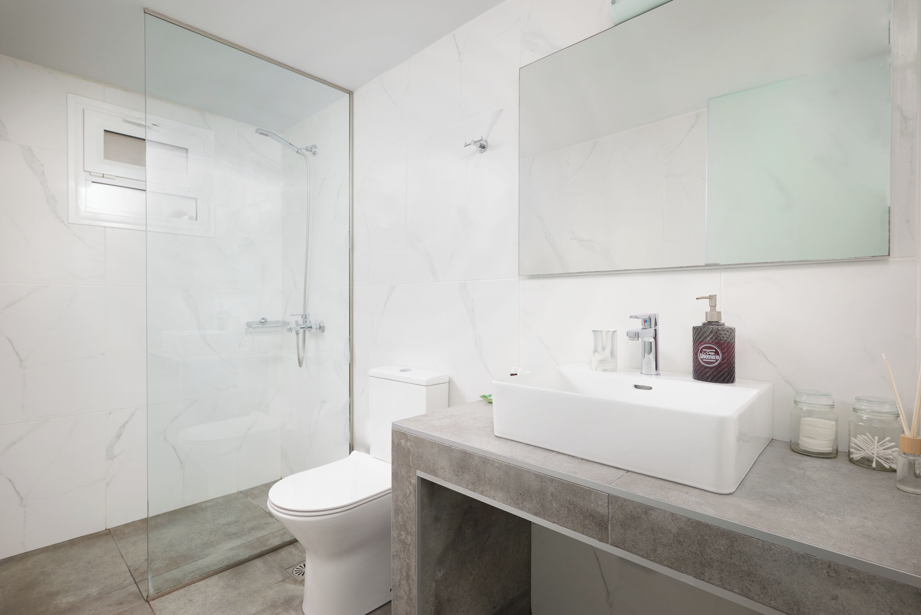 Een moderne badkamer met een douchecabine van glas, een wit toilet en een grote spiegel boven een witte wastafel met een marmeren aanrechtblad.