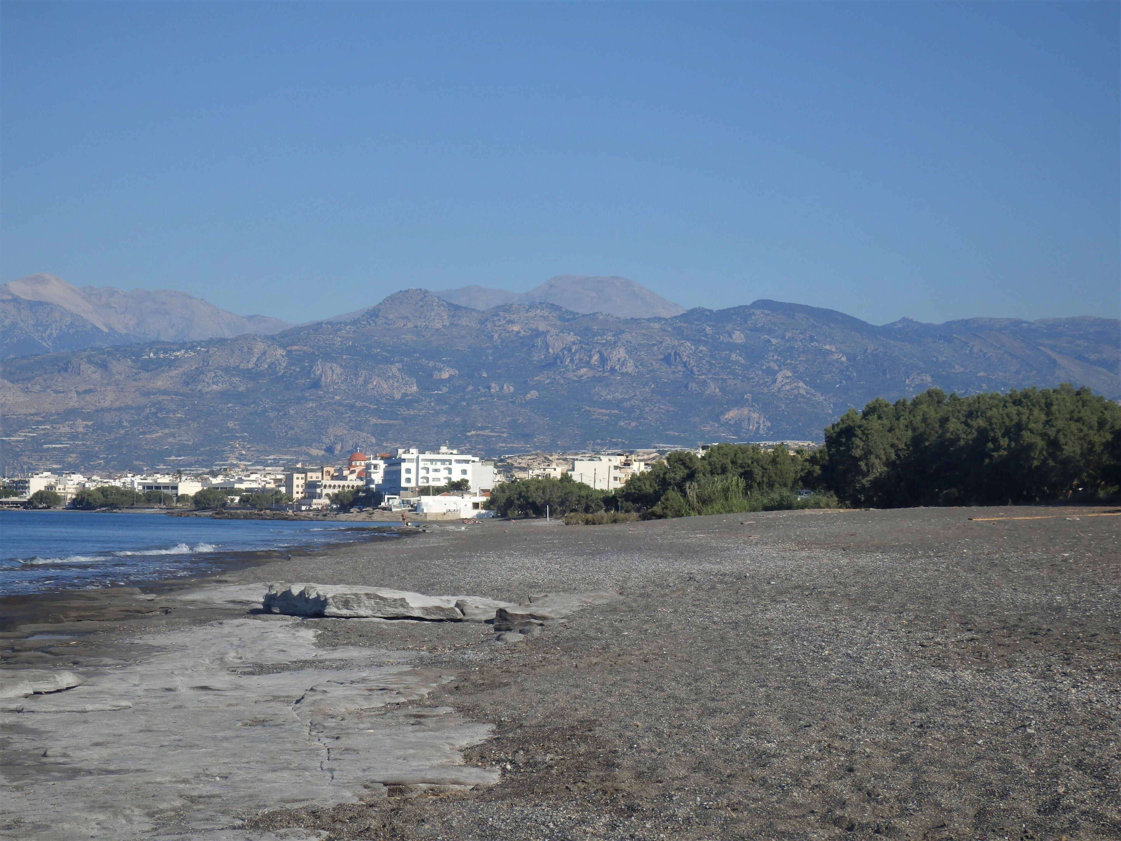 A rocky beach with buildings in the background and mountains in the distance.