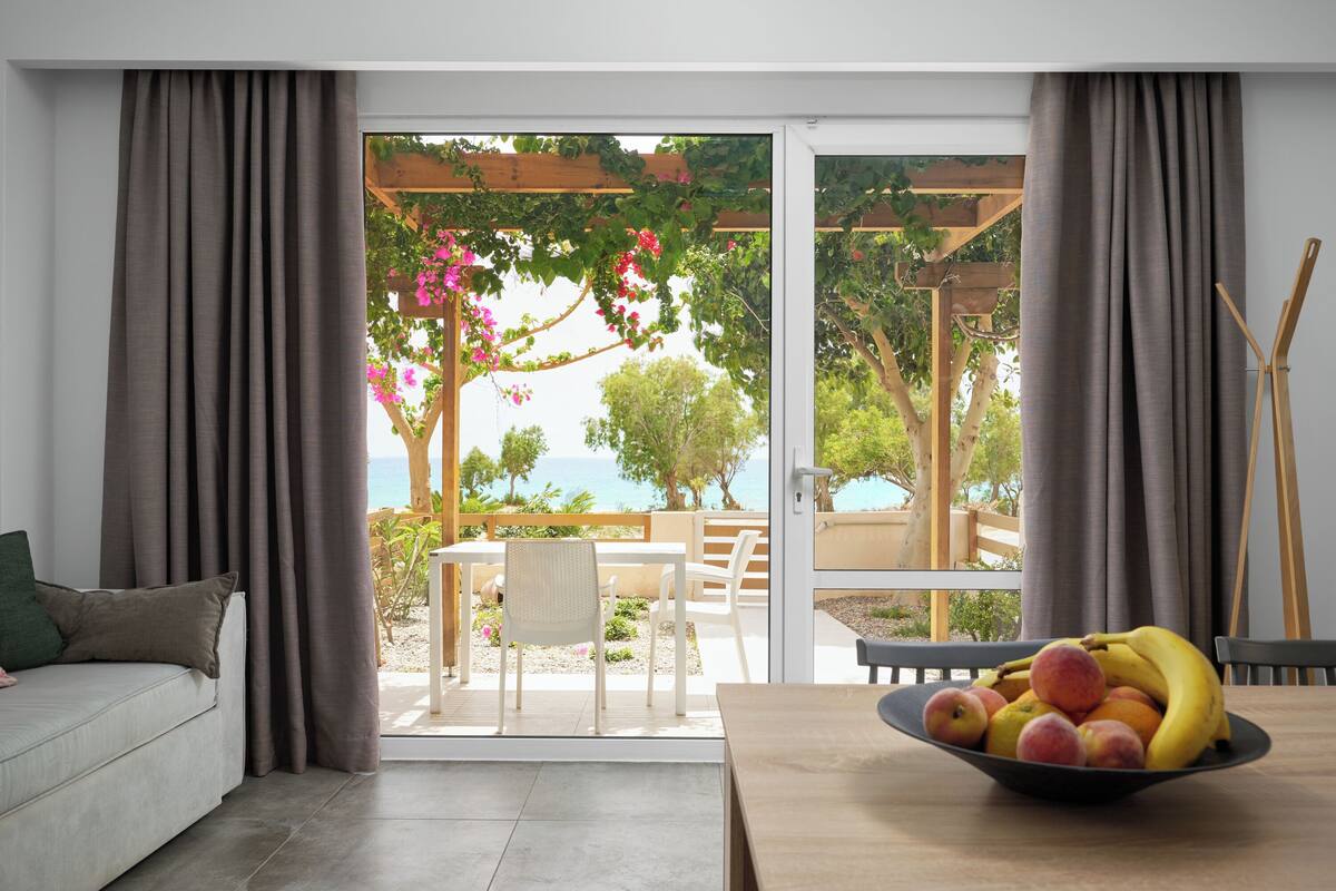 Une pièce avec vue sur la mer, une table en bois sur laquelle se trouve un panier de fruits, un canapé et un coin repas avec des chaises.