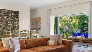 Een moderne woonkamer met een bruine modulaire bank, decoratieve wandpanelen en uitzicht op een tropische tuin door grote ramen.