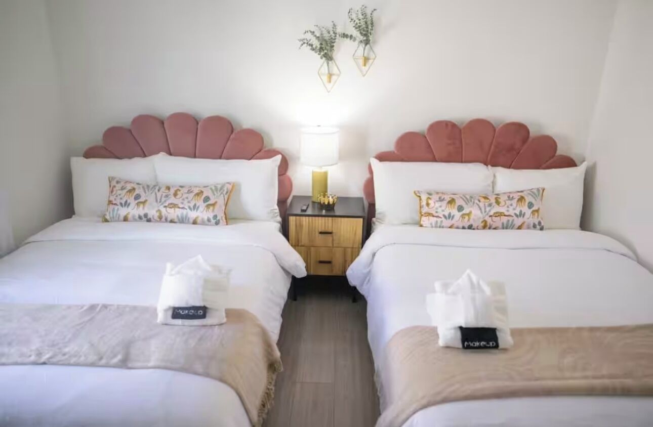 Une chambre d’hôtel avec deux lits, chacun doté d’une tête de lit rose, de draps blancs et de coussins décoratifs. Une table de chevet avec une lampe et une petite plante se trouve entre les deux lits.