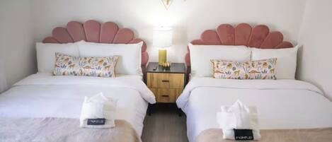 A hotel room with two beds, each with a pink headboard, white bedding, and decorative pillows. There is a nightstand with a lamp and a small plant between the beds.