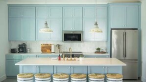 Une cuisine avec des meubles de couleur bleu clair, un réfrigérateur en acier inoxydable, un îlot central doté de tabourets et des luminaires suspendus.
