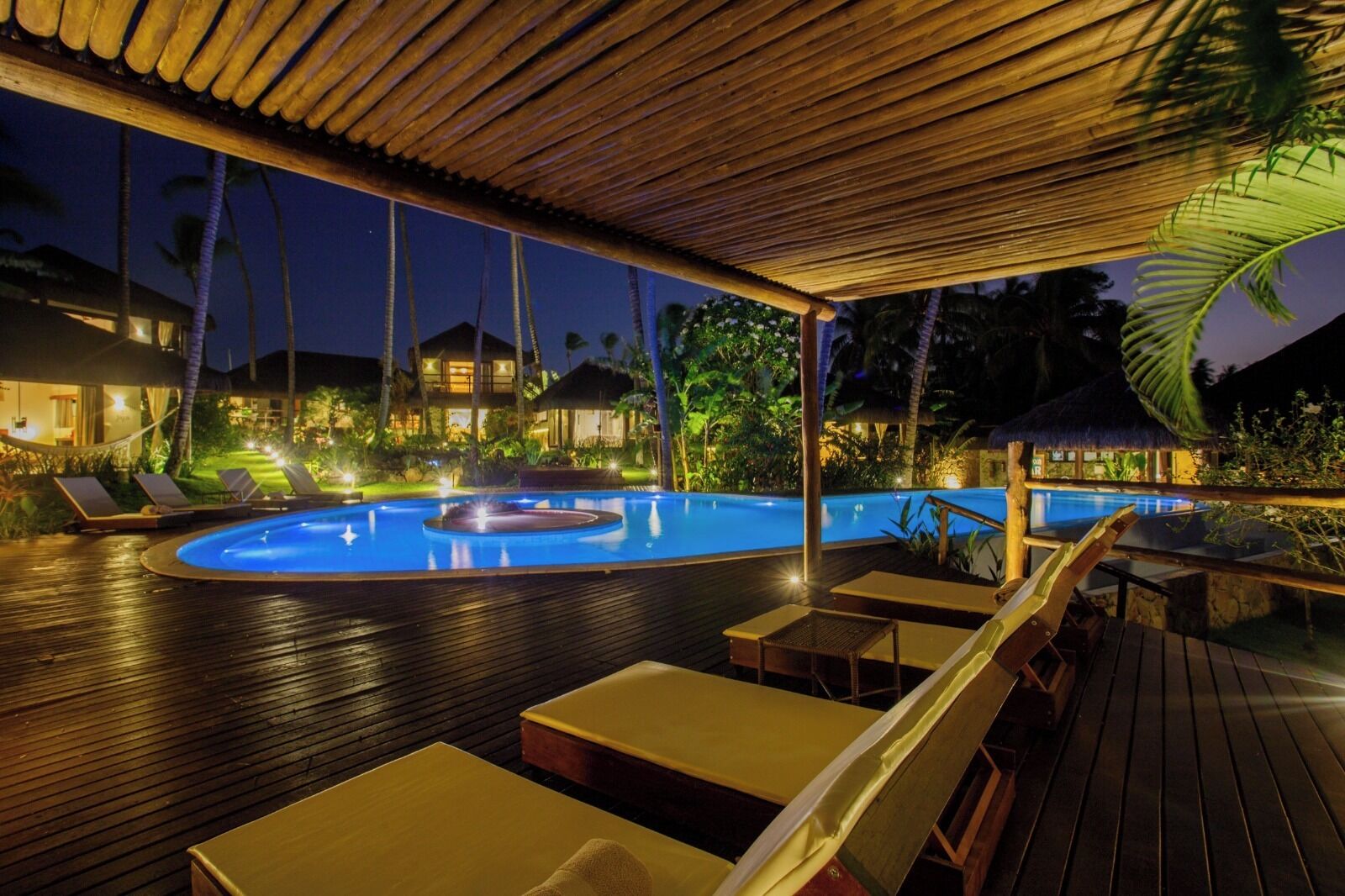 Un espace piscine nocturne avec des chaises longues et un jacuzzi, entouré d’une terrasse en bois et de palmiers.