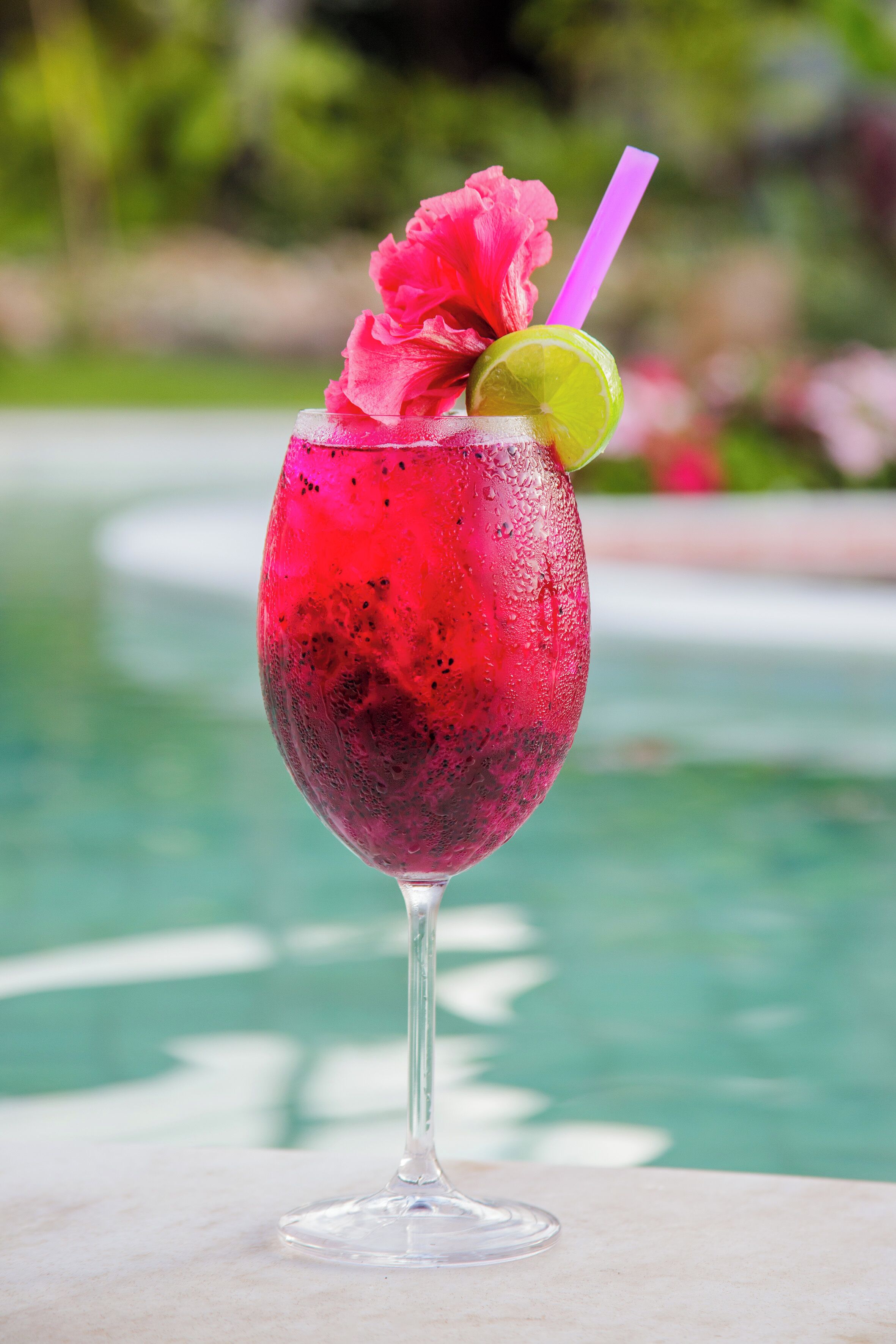 Un verre à cocktail contenant une boisson rose, un quartier de citron vert et une fleur, placé près d’une piscine.