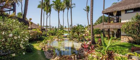 A tropical garden with a pond, palm trees, and thatched-roof structures.