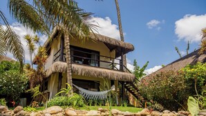 A two-story building with a thatched roof, surrounded by a tropical garden and a pond. - Pousada Jaya Boutique (São Miguel do Gostoso)