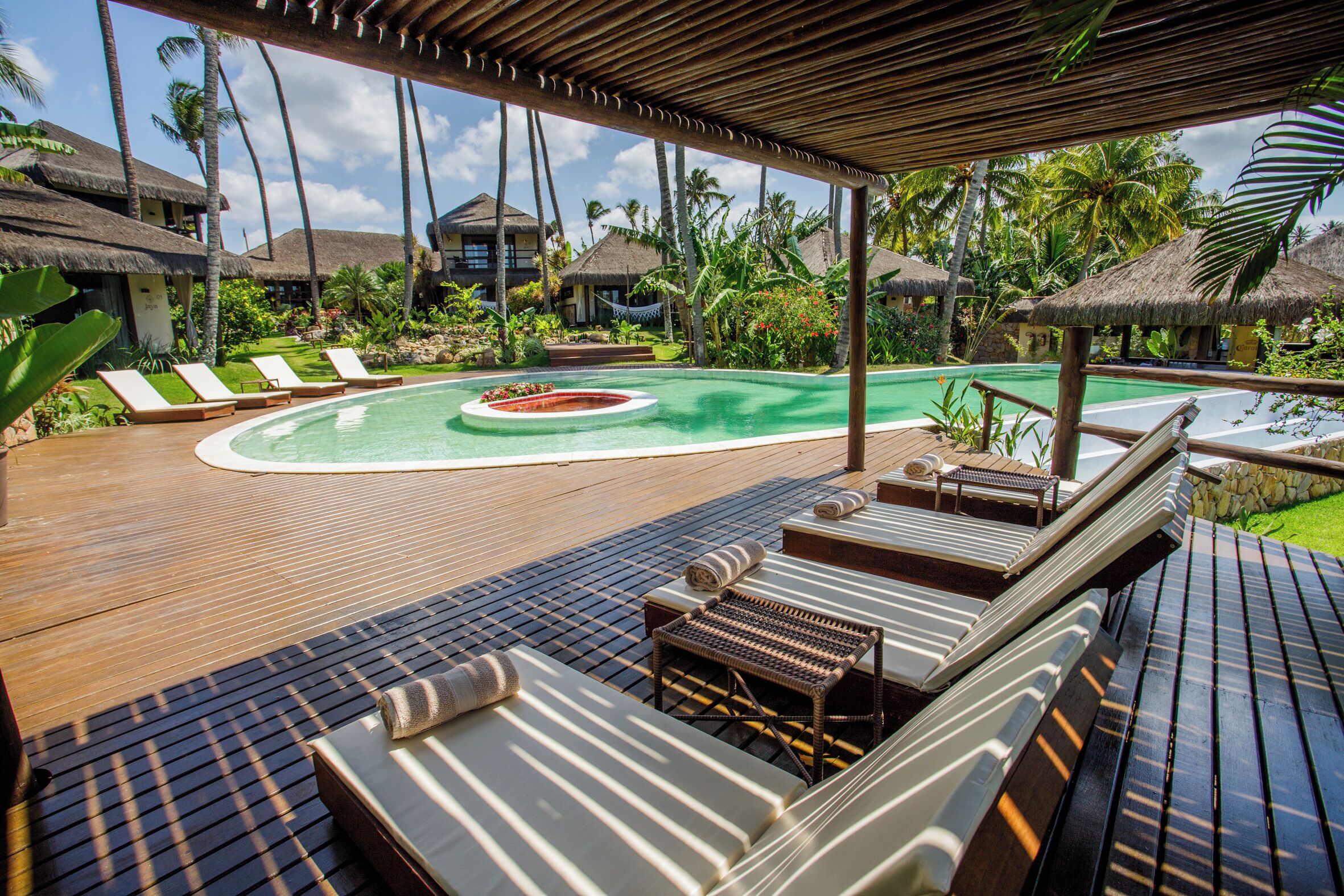 Una terraza de madera con sillones de descanso y una pequeña zona de piscina, rodeada de vegetación tropical y estructuras de techo de paja.