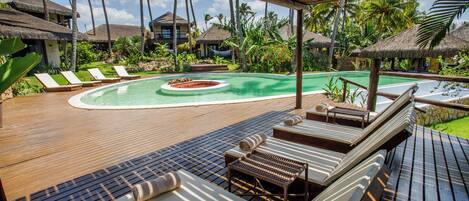 Una terraza de madera con sillones de descanso y una pequeña zona de piscina, rodeada de vegetación tropical y estructuras de techo de paja.