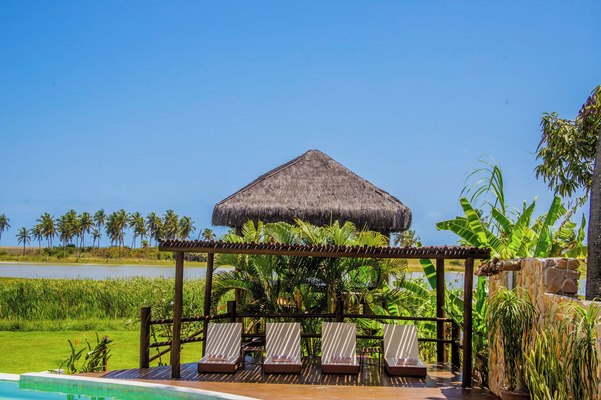 A poolside area with lounge chairs, a thatched-roof gazebo, and lush greenery.
