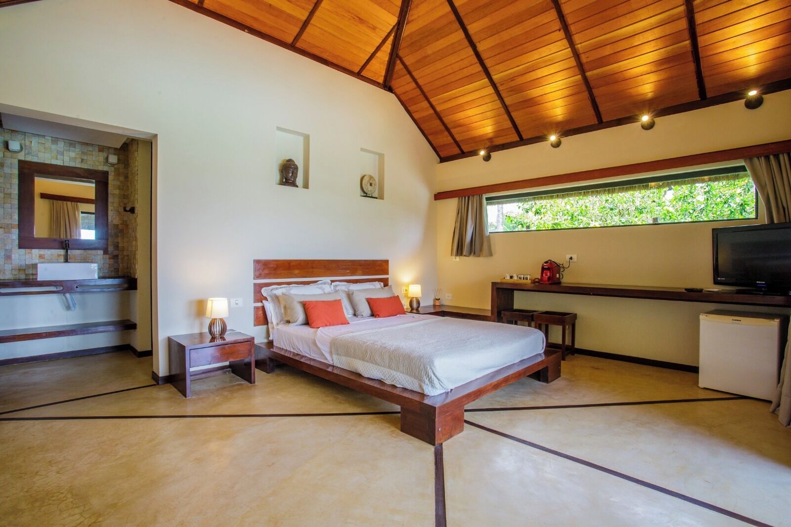 A bedroom with a wooden ceiling, a bed with white bedding and red pillows, a desk with a television, and a window with a view of greenery.