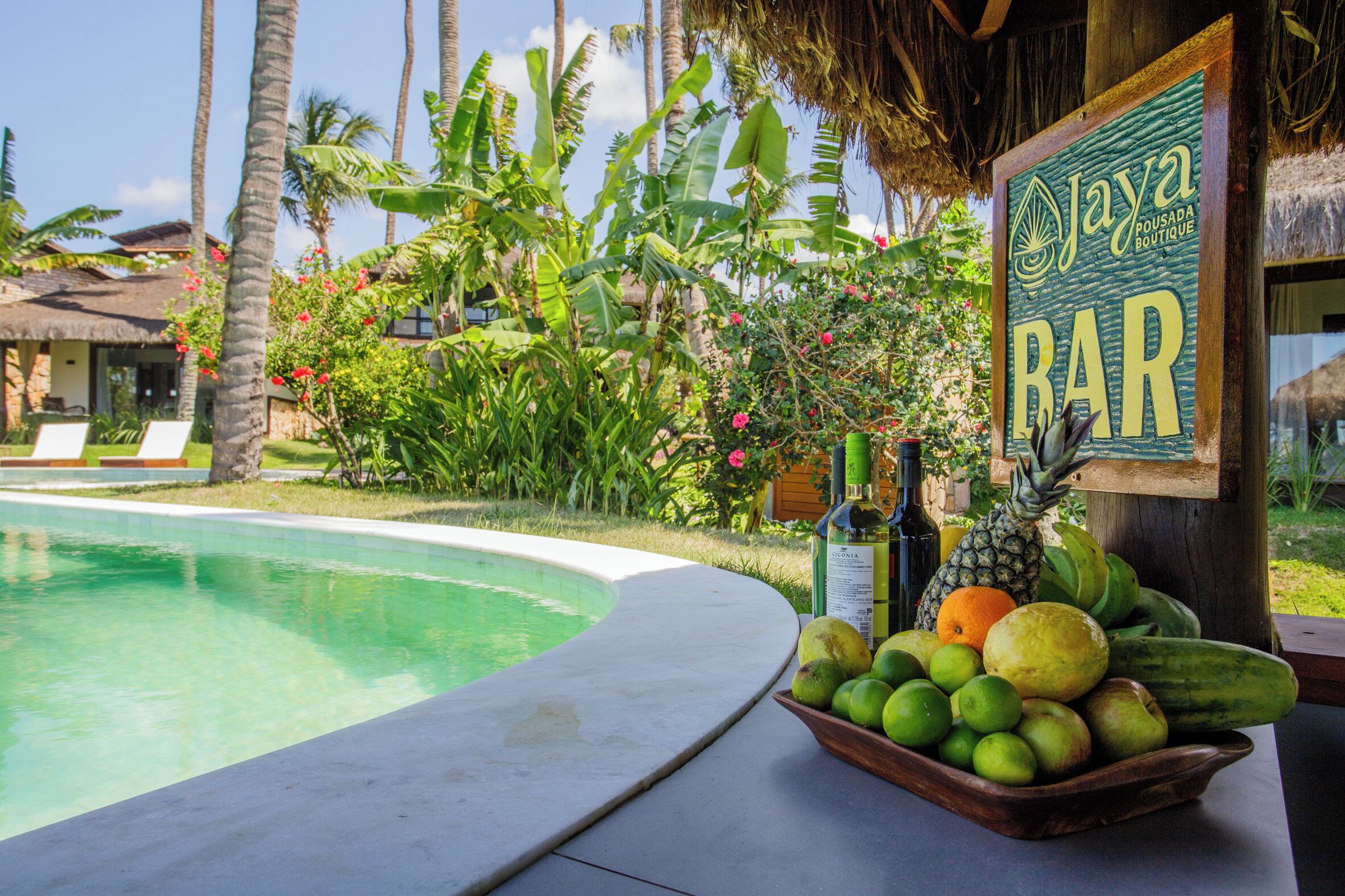 A poolside area with a bar sign, a bowl of fruit, and tropical vegetation.