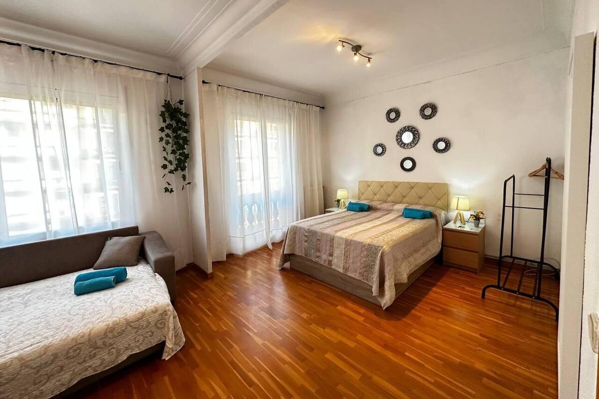 A bedroom with two beds, wooden flooring, a bedside table with a lamp, and a hanging plant.