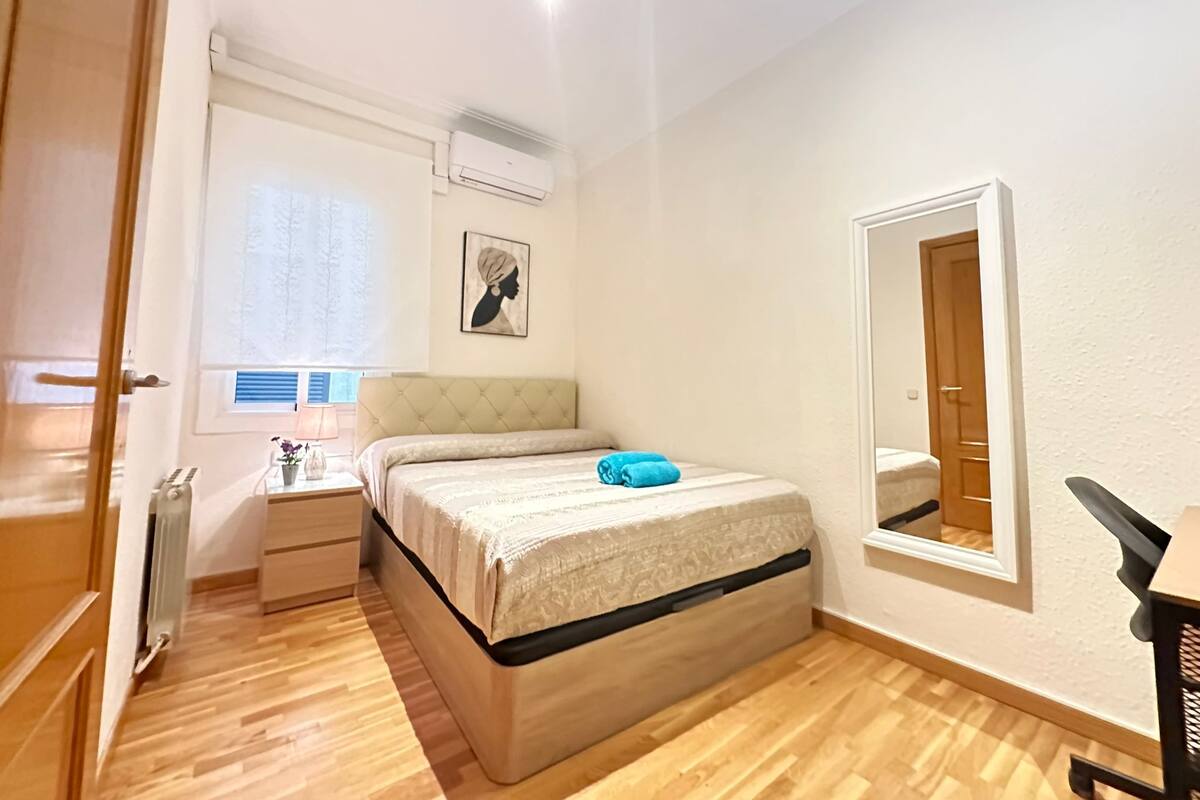 A bedroom with a bed, bedside table, lamp, window, air conditioning unit, and a mirror.