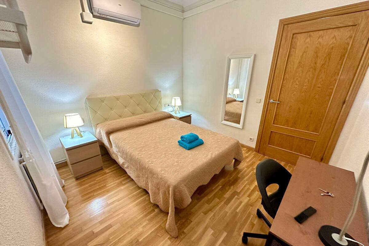 A bedroom with a bed, bedside tables, a desk, a chair, a mirror, and a door.